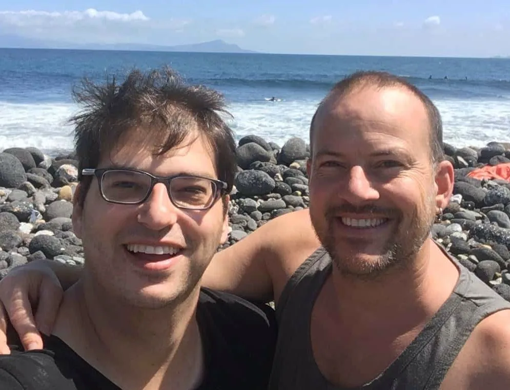 Micah and David smiling together on a rocky beach in San Miguel, the ocean behind them.