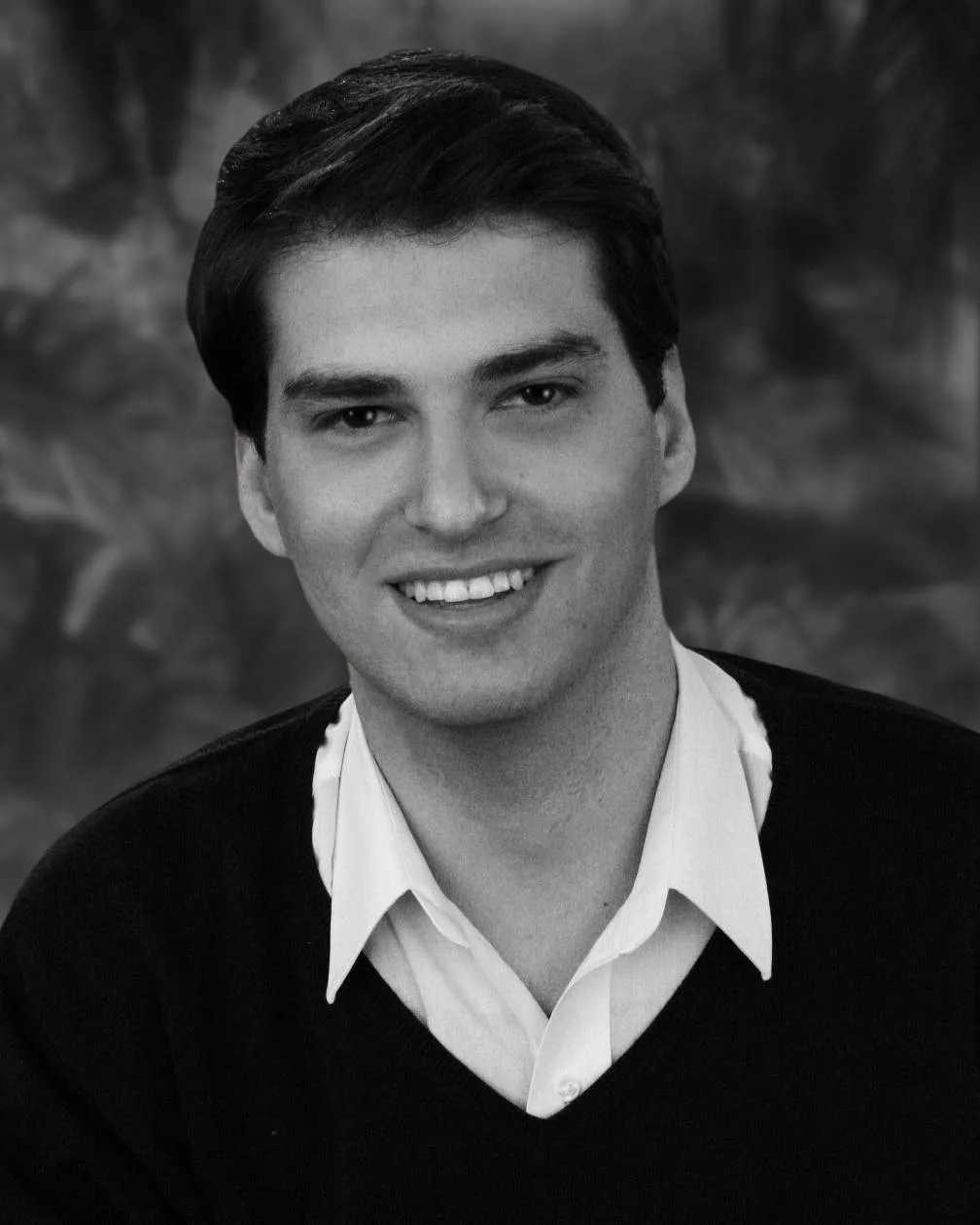 Black-and-white portrait of Micah Bleich, smiling, in a dark sweater over a white collared shirt.